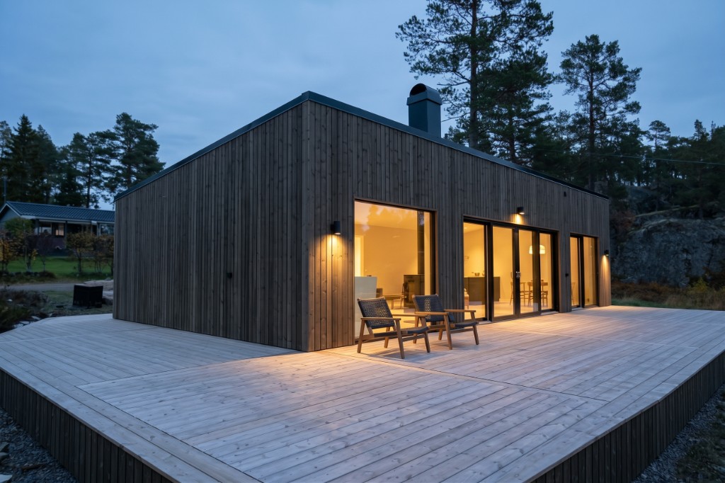Ett mörkt trähus i blå timmen, glasdörrar mot en ljus träaltan, tallar och berg bakom.