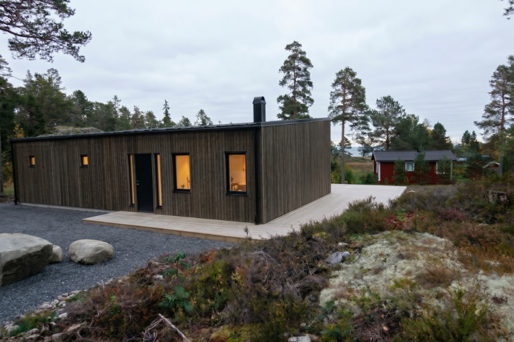 Framsidan av trähuset bland tallar, med lav, ljung och sten i förgrunden.